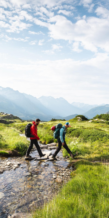 Due escursionisti attraversano un piccolo ruscello sul Sentiero Alpino Calanca, circondati dal paesaggio alpino
