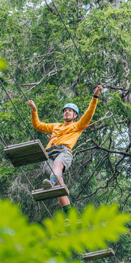 Persona con casco e imbragatura in equilibrio su un ponte sospeso nel parco avventura San Bernardino tra gli alberi