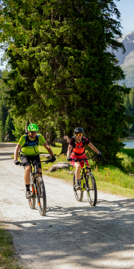 Zwei Personen auf Mountainbikes beim Lago d’Isola in San Bernardino, umgeben von Wald und Bergen