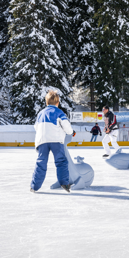Kinder und Erwachsene fahren auf der Kunsteisbahn in San Bernardino Schlittschuh, umgeben von verschneiten Bäumen und Wintersonne