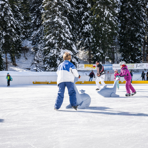 Kinder und Erwachsene fahren auf der Kunsteisbahn in San Bernardino Schlittschuh, umgeben von verschneiten Bäumen und Wintersonne