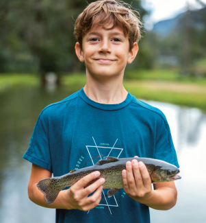 Ad Augio, presso la Pesca Sportiva, si pesca il proprio pranzo (Foto: © Nico Schaerer) Un ragazzo tiene con orgoglio una trota fario appena pescata con entrambe le mani mentre sorride alla telecamera