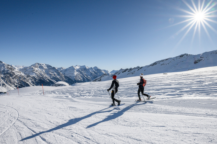 Zwei Personen beim Schneeschuhwandern auf einer verschneiten Hochebene mit Bergpanorama und Sonnenschein