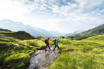 Zwei Wanderer überqueren einen kleinen Bach auf dem Sentiero Alpino Calanca inmitten einer alpinen Berglandschaft
