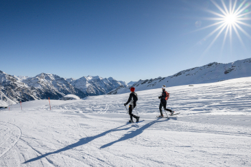 Zwei Personen beim Schneeschuhwandern auf einer verschneiten Hochebene mit Bergpanorama und Sonnenschein