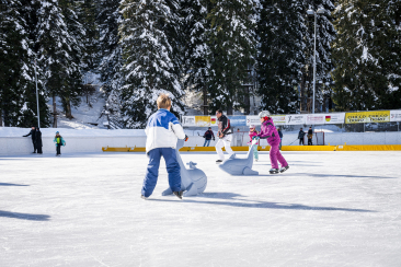 Bambini e adulti pattinano sulla pista di ghiaccio di San Bernardino, circondati da alberi innevati e dal sole invernale